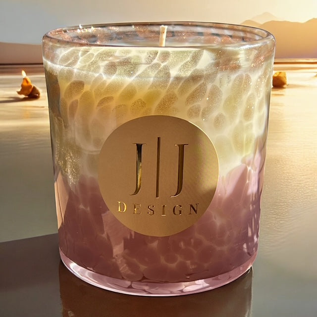 Candle with a gold logo and 'JJ Design' text on a reflective surface with a scenic background.