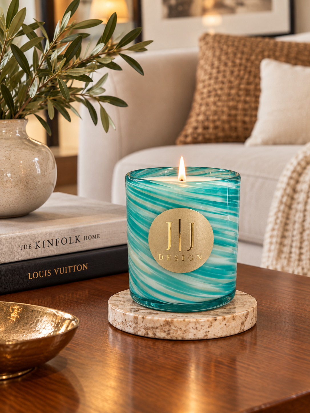 A teal and turquoise marbled glass jar candle with a gold circular logo on the front, lit and placed on a round stone coaster on a wooden table in a cozy living room setting.